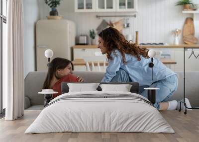 Cheerful energetic woman tickles teenage girl to cheer and make child laugh. Happy close-knit family of mother and daughter spend time together during school holidays having fun at sofa in apartment  Wall mural