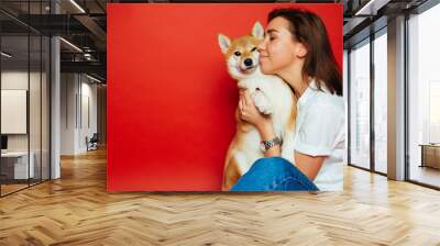 Cute brunette woman in white t shirt and jeans holding and embracing Shiba Inu dog on plane red background. Love to the animals, pets concept Wall mural