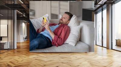 Disinterested man with phone lying on couch surfing online web, procrastination at home. Lazy male with social media addiction on smartphone scrolling internet, enjoy virtual life rest look at screen. Wall mural