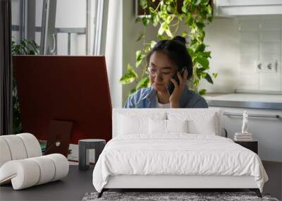 Enterprising pensive Asian woman sits at computer desk in home office making phone call. Professional casual Chinese girl in search of work calls companies wanting to get job as secretary or clerk Wall mural