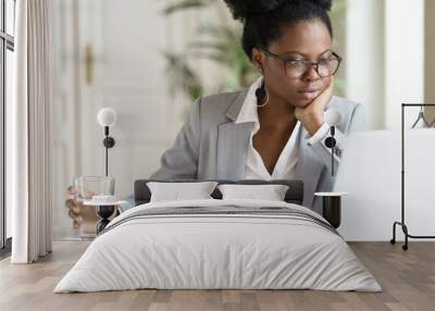 Focused young African American businesswoman or student in blazer wear glasses working at laptop at home office, holding glass of water, looking at screen computer, reading interesting article online. Wall mural