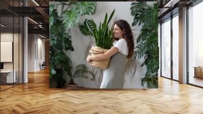 Home gardening. Young happy pleased italian woman gardener in dress holding Sansevieria houseplant in wicker planter while working in home garden full of green lush tropical indoor plants Wall mural