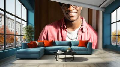 Indoor portrait of happy optimistic young African American man wearing glasses smiling at camera, standing by window in sunny room at home. Positive attitude and emotional wellbeing concept Wall mural