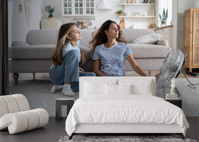 Loving carefree mother and teenage girl sits in relaxed position on floor in living room near fan. Satisfied positive family of woman and daughter spend time at home during abnormally high temperature Wall mural