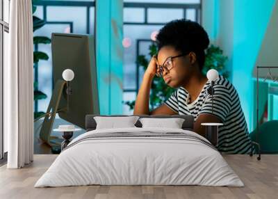 Pensive African woman wearing glasses works freelancer sitting at table at home. Kinky-haired entrepreneur thinks about new business project looking at monitor of computer against potplants by window Wall mural