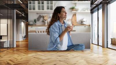 Pleased smiling woman resting on sofa with cup of tea enjoying alone time at home, looking aside with dreamy face expression. Positive female starting day with morning coffee. Free time for parent Wall mural