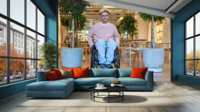 Relaxed confident disabled man in wheelchair leaving cafe, sitting between two potted trees, facing camera. Accessible public space encourages active lifestyle, provides comfort in outdoor areas Wall mural