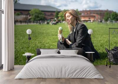 Satisfied girl resting on grass, snacking on yummy sundae in waffle cone, bathing in bright warm rays of summer sun. Female student in suit sitting on campus eating dessert lunch between classes Wall mural