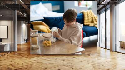 Smiling child schoolboy pouring milk into glass while having healthy breakfast at home, happy kid boy sitting at kitchen table eating cereals. School-aged children and morning meal Wall mural