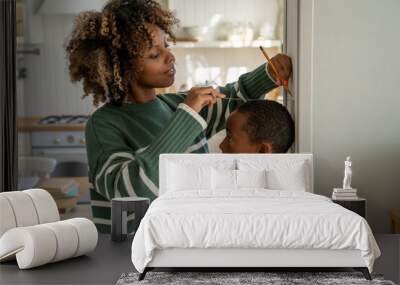 Smiling loving African American mother measuring height of child son at home, using metal ruler and pencil, marking top of head on wall, mom taking measurement while boy standing against flat surface Wall mural