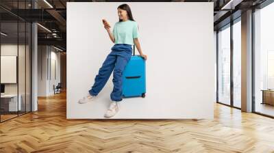 Smiling teenage girl sits on travel blue suitcase and using smartphone. Happy teen girl tourist inspired by upcoming trip chooses route on phone isolated on gray studio background with copy space Wall mural