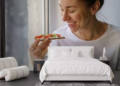 Smiling woman eating rye crisp bread with creamy vegetarian cheese tofu, cherry tomato and rucola micro greens, sitting at home and looking at window. Healthy food, gluten free, diet concept.  Wall mural