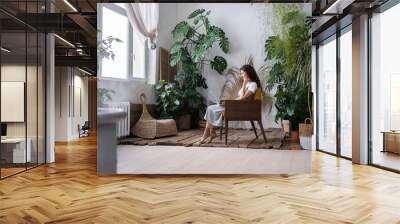 Smiling young female owner city greenhouse sitting on chair in front of window with laptop having rest, buying plants online, looking intresting serias, texting with friends. Monstera on background. Wall mural