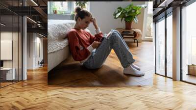 Teens and cyberbullying. Upset teen girl sitting on floor near bed using smartphone at home, scrolling social media. Child spending too much time on phone. Teenagers and gadget addiction Wall mural