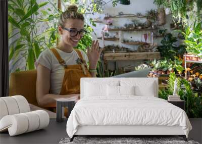 Woman gardener wear orange overalls using laptop after work, smiling and talking in video chat, meeting her friends, sitting on chair in greenhouse surrounded by exotic plants. Home gardening.  Wall mural