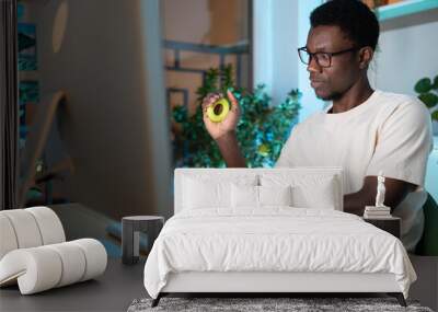 Young African man freelancer exercising with silicone grip ring at cozy home office, remote worker doing hand exercises while resting from computer at workplace. Carpal tunnel syndrome prevention Wall mural