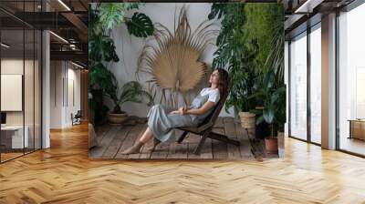 Young calm hipster woman relaxing sit on comfortable armchair in stylish scandinavian indoor garden with green exotic plants. Happy florist girl enjoy spending time with houseplant in home greenhouse Wall mural