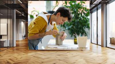 Young female artisan concentrated on modeling jug from raw clay while pottery lesson or workshop in creative studio. Woman ceramic business owner making craft for sale in handmade potter retail store Wall mural