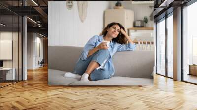 Young happy relaxed Latin woman enjoying quiet moments at home with cup of tea or coffee, smiling female holding mug with hot drink resting on sofa in living room and dreaming, relaxing of everything Wall mural