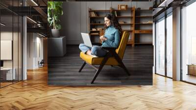 Young woman freelancer sitting with crossed legs on chair at comfortable remote workplace, consulting remotely from home, female consultant in wireless headset having video chat with client via laptop Wall mural