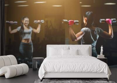 Slim, bodybuilder girl, lifts heavy dumbbell standing in front of the mirror while training in the gym. Sports concept, fat burning and a healthy lifestyle. Wall mural