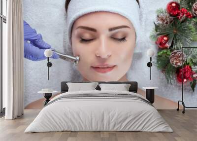 The cosmetologist makes the procedure Microdermabrasion of the facial skin of a beautiful, young woman in a beauty salon.Closeup woman face and new year wreath. Wall mural