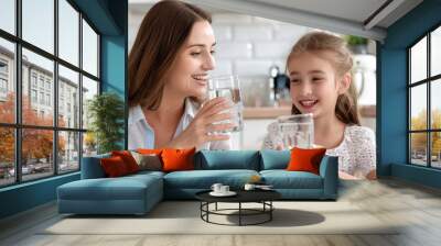 Young beautiful mother and little girl drinking fresh pure water from glasses in kitchen Happy mom and daughter holding glasses with water. Eating habits, water filter advertisement, healthy nutrition Wall mural