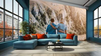 Rear view of couple in love who standing on mountain, watching around beautiful mountain scenery and tenderly embracing each other. Pleasant moment of hugs while walking in the fresh air. Wall mural