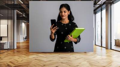 Pretty young girl holding book and using mobile on grey background Wall mural