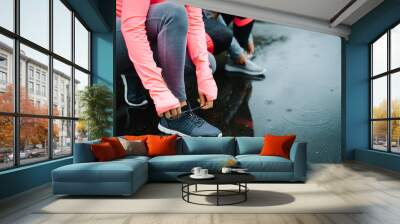 Urban athletes lacing sport footwear for running over asphalt under the rain. Two women getting ready for outdoor training and fitness exercising on cold winter weather. Wall mural