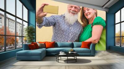 Trendy senior couple taking a selfie outdoor with yellow wall backgrou - 60's people having fun with new technology trends - Concept of joyful elderly and travel - Main focus on woman face Wall mural