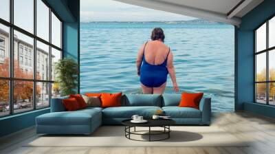 Rear view of overweight woman in blue swimsuit standing in still sea waters. Plus size woman at beach. Wall mural