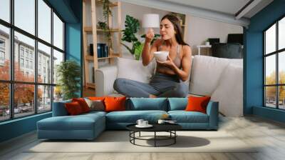 Beautiful young woman is sitting on the sofa in the living room and eating oatmeal Wall mural