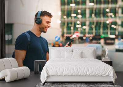 Young sport man walking around in the city and listens to music via smartphone through wireless headphones Wall mural