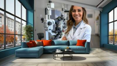 A smiling female eye doctor with crossed arms wearing a lab coat standing in her well equipped ophthalmology clinic Wall mural