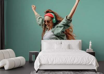 A woman in casual attire and headphones showcasing a playful dance move against a soft green backdrop Wall mural