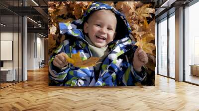 Laughing little blond baby in warm jacket lies on fallen golden autumn leaves on ground and happy. Autumn childrens fun to lie in colorful fallen leaves outdoor sunny day. Close up portrait of kid Wall mural