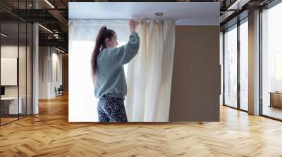 Young tender girl broadcasts curtains on a special fastening near the window Wall mural