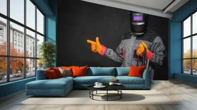 Welder man in the helmet with a welding terminals is showing by the index finger on the copy space on the black background. Wall mural