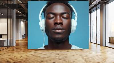 A young man with headphones enjoys a peaceful moment, fully absorbed in the music as he relaxes against a soothing blue background Wall mural