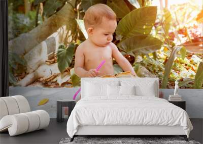 Pensive and passionate infant baby sits on the ground and eats and drinks green young coconut with spoon and drinking straw Wall mural