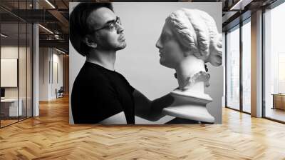 Black and white portrait of adult man sculptor, artist in black t-shirt and glasses holding in hands, looking at antique sculpture woman head over light gray background Wall mural