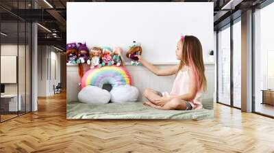 Cheerful pretty small girl with decorative bow and pink strands in hair sitting on bed and playing with collection of dolls, side view. Happy childhood, cheerful lifestyle, games, comfortable pastime Wall mural