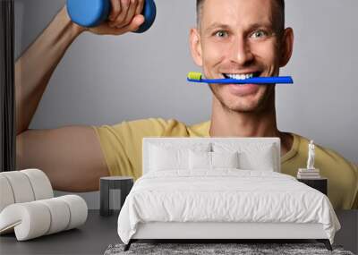 Ecstatic young adult man in yellow t-shirt holding blue toothbrush in his mouth, teeth and dumbbell in hand. working out, doing morning exercises and activities. Healthy vigorous lifestyle concept Wall mural