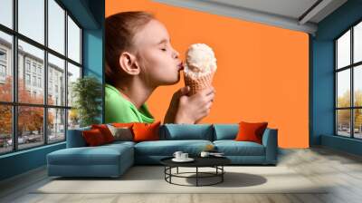 Little baby girl kid kissing vanilla ice cream in waffles cone on yellow orange background in green t-shirt Wall mural