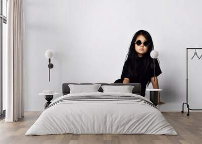 Pretty self-confident early adult serious asian kid girl in stylish black dress, white sneakers and sunglasses sits on floor at copy space over white wall background. Trendy children fashion concept Wall mural