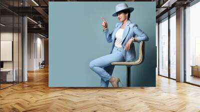 Slyly smiling short haired brunette woman in blue business smart casual suit and hat sitting on stool pointing finger at copy space over blue background. Stylish female wear and fashion concept Wall mural