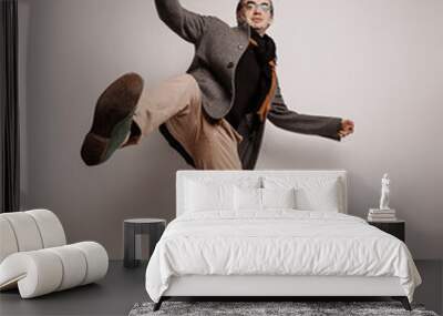 Stylish adult man hipster in pants and plaid jacket stands holding foot up, making giant step, has lost balance, falling down waving hands over grey background Wall mural