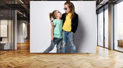 Two frolic kid girls sisters in stylish casual clothing, sunglasses and sneakers stand together and look at each other Wall mural