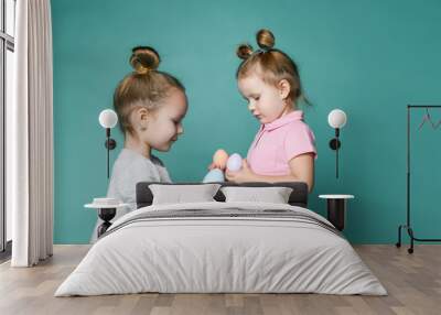 Two small happy girls in cosy home clothing standing face to face and holding pastel colored eggs in hands Wall mural